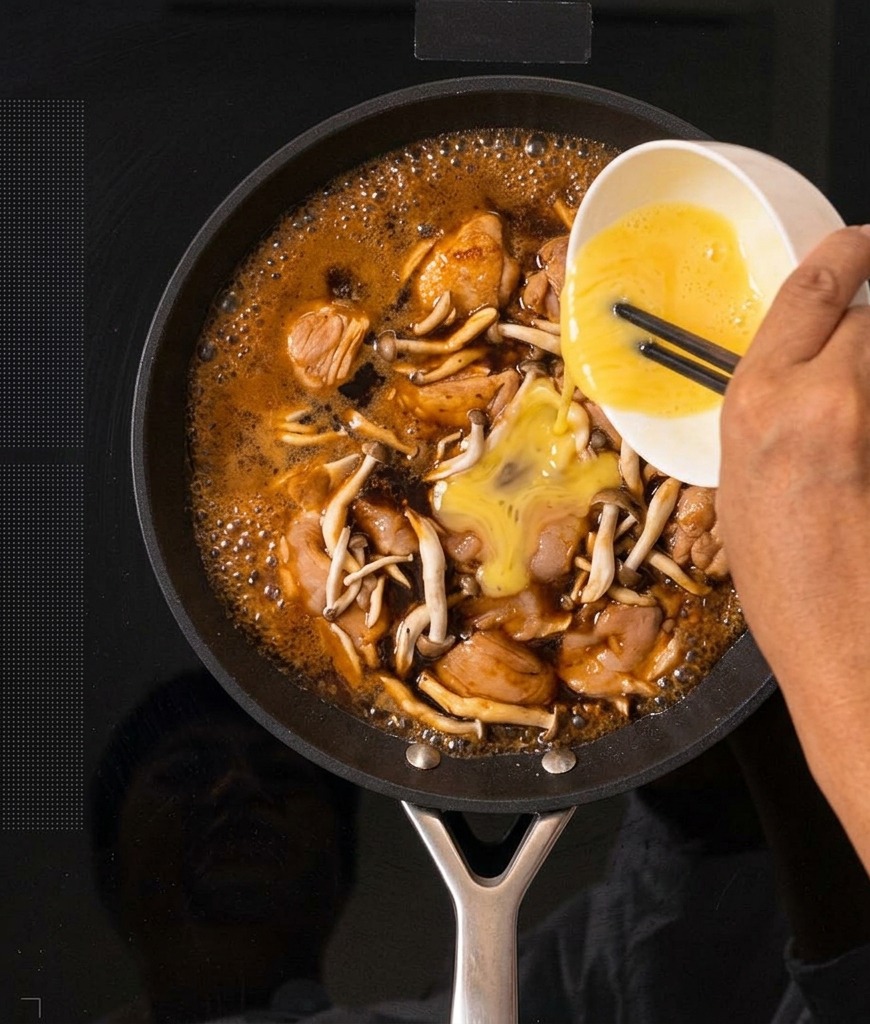 Uova sbattute di colore giallo versate da una ciotola bianca in una padella con il composto di Oyakodon che bolle.