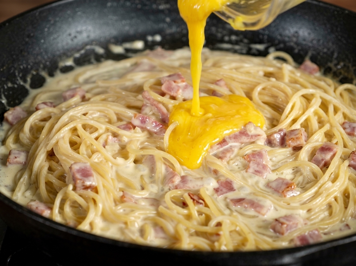 Crema gialla di tuorlo e parmigiano versata sugli spaghetti cremosi in padella.