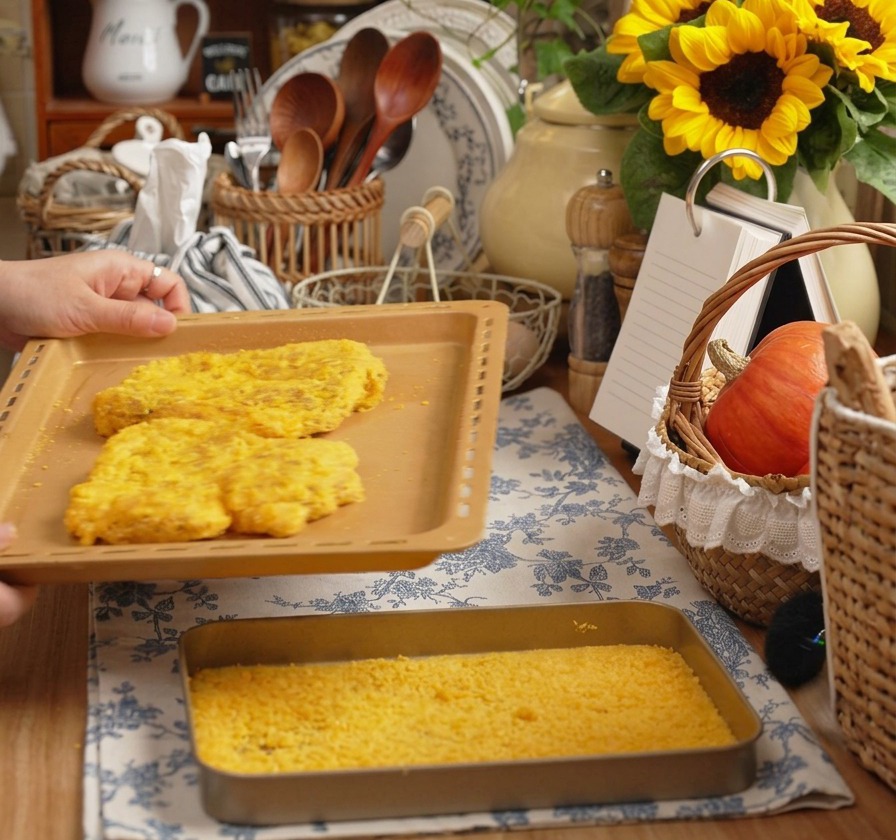 Mani che tengono una teglia con due grandi cotolette di maiale impanate in una cucina accogliente.