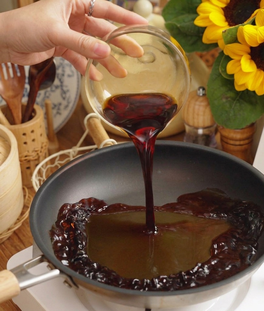 Salsa scura e saporita versata da una ciotola di vetro in una padella nera su un piano di cottura elettrico bianco.