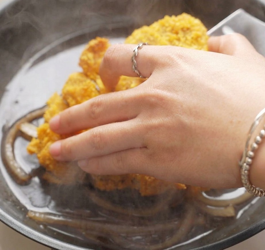Una mano che adagia fette di cotoletta di maiale impanata e dorata in una padella piena di cipolle affettate che sobbollono in una salsa scura saporita.
