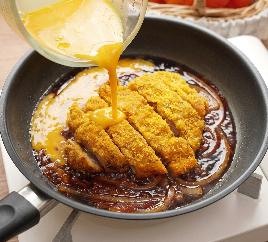 Uova sbattute gialle versate da un misurino di vetro in una padella contenente una cotoletta di maiale fritta a fette e cipolle.