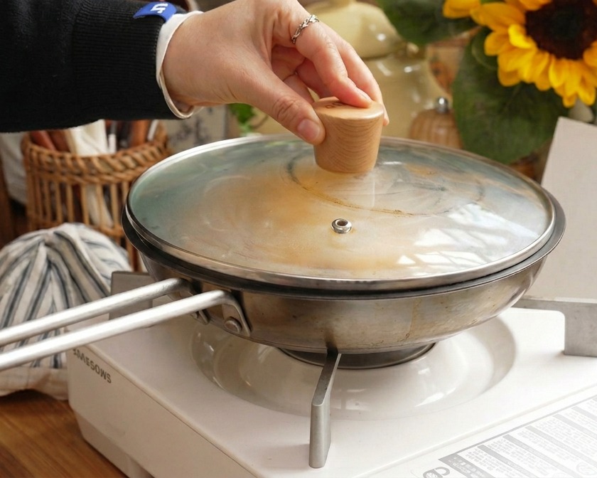 Una mano che tiene il pomello di legno di un coperchio di vetro mentre viene posizionato su una padella in acciaio inossidabile per cuocere a vapore il piatto di maiale e uova.