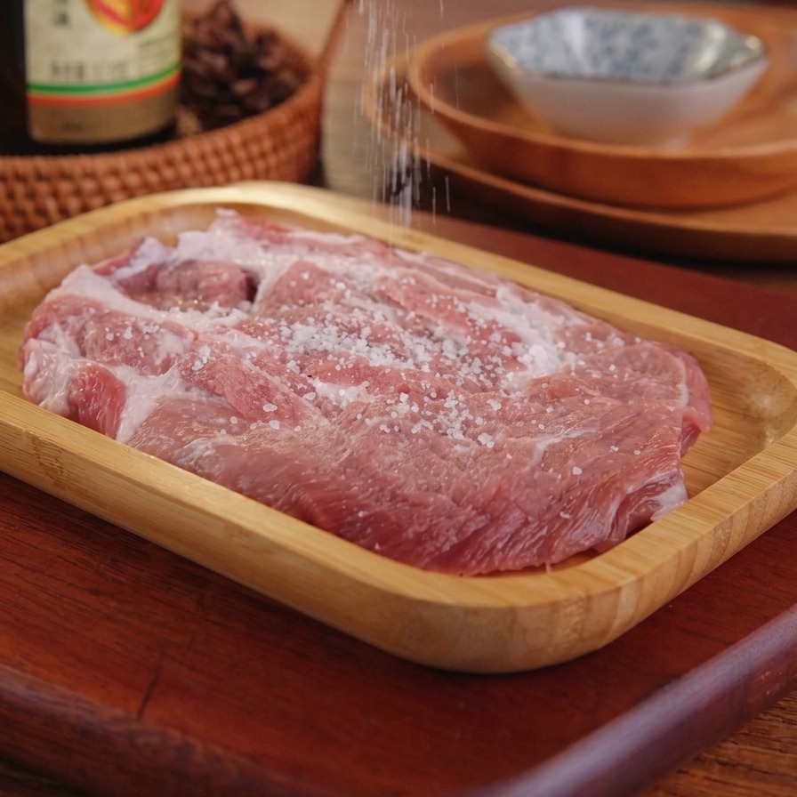 Sale che viene cosparso con la mano su una cotoletta di maiale cruda adagiata su un vassoio di legno.