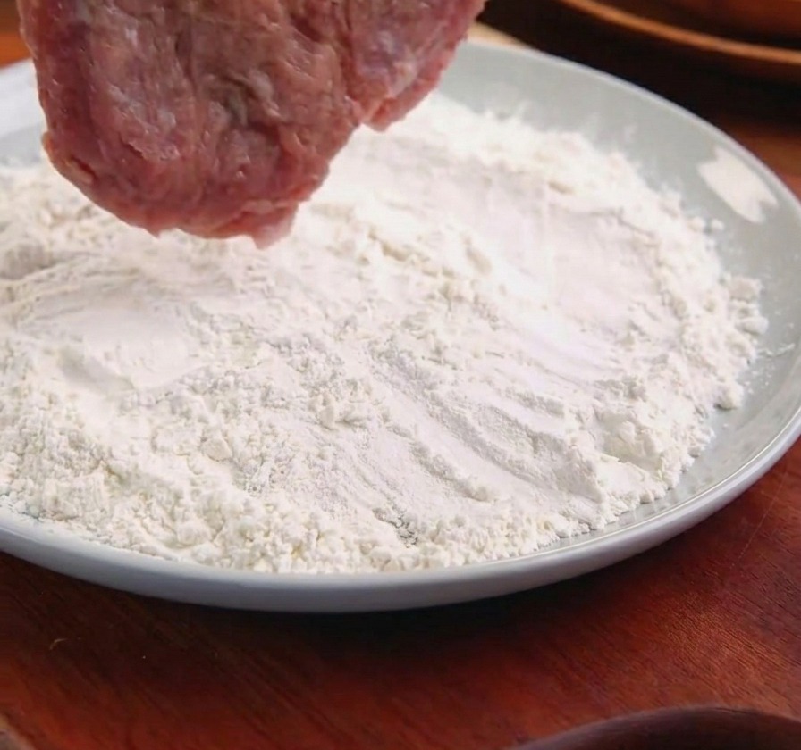 Una cotoletta di maiale tenuta da pinze di metallo mentre viene passata in un piatto di farina bianca.