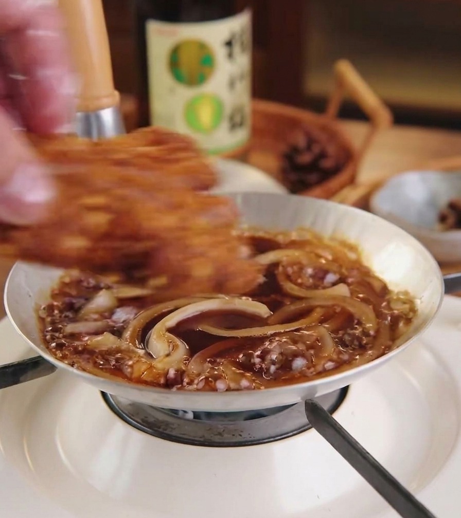 Una cotoletta di maiale fritta e dorata posta sopra uno strato di cipolle stufate e salsa in una piccola padella.