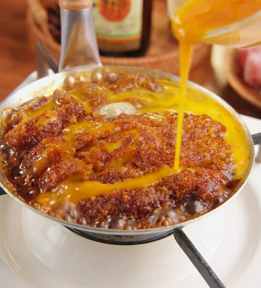 Uovo giallo sbattuto che viene versato su una cotoletta di maiale impanata che sobbolle in una salsa scura con cipolle.