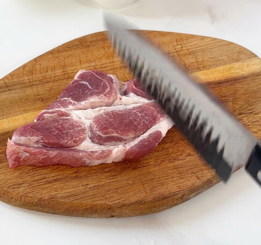 Una mano che utilizza il dorso di un coltello da chef per battere una fetta di maiale cruda su un tagliere di legno.