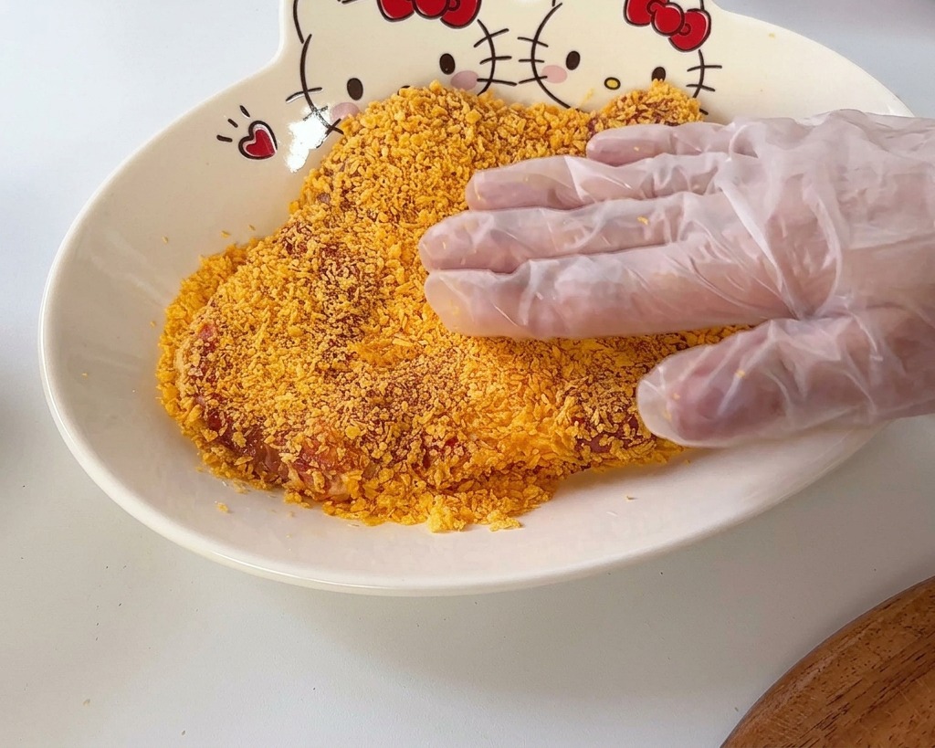 Una mano che preme una cotoletta di maiale su uno strato di pangrattato arancione in una ciotola con un design di Hello Kitty.