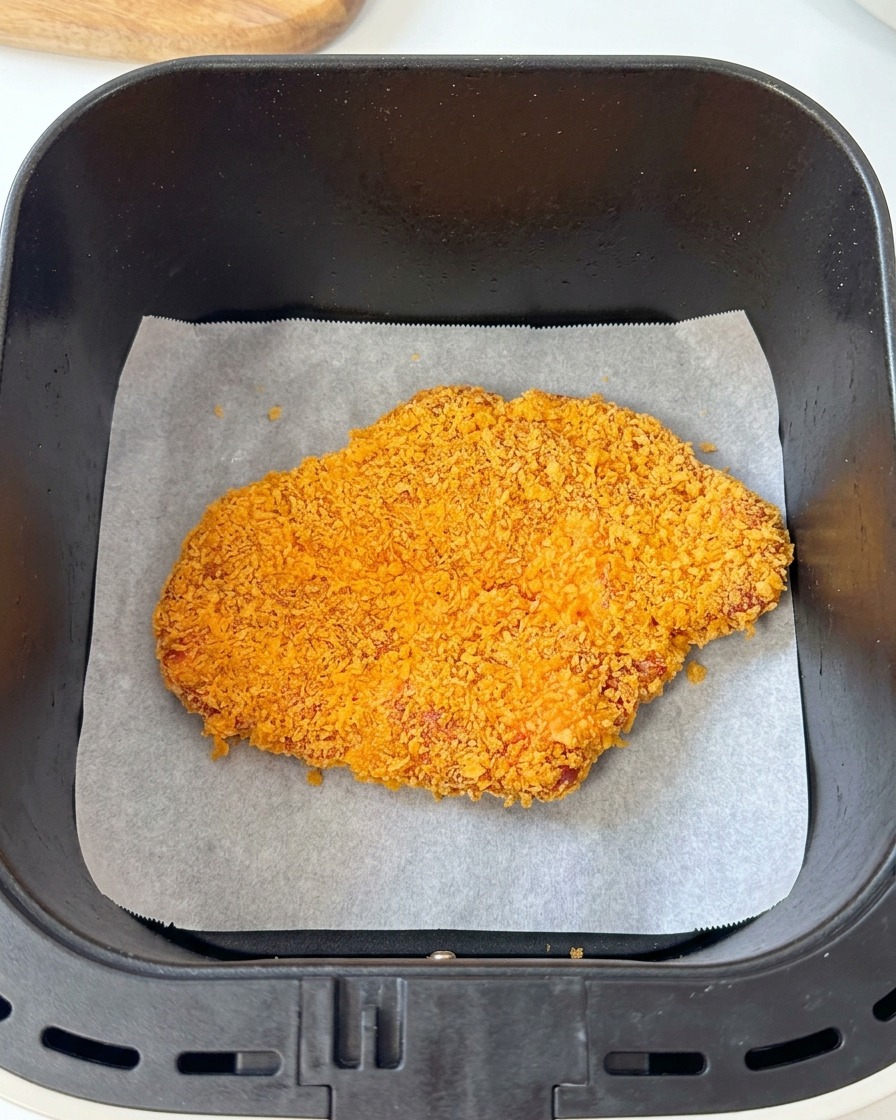 Una cotoletta di maiale impanata che riposa su carta da forno allinterno del cestello nero di una friggitrice ad aria.