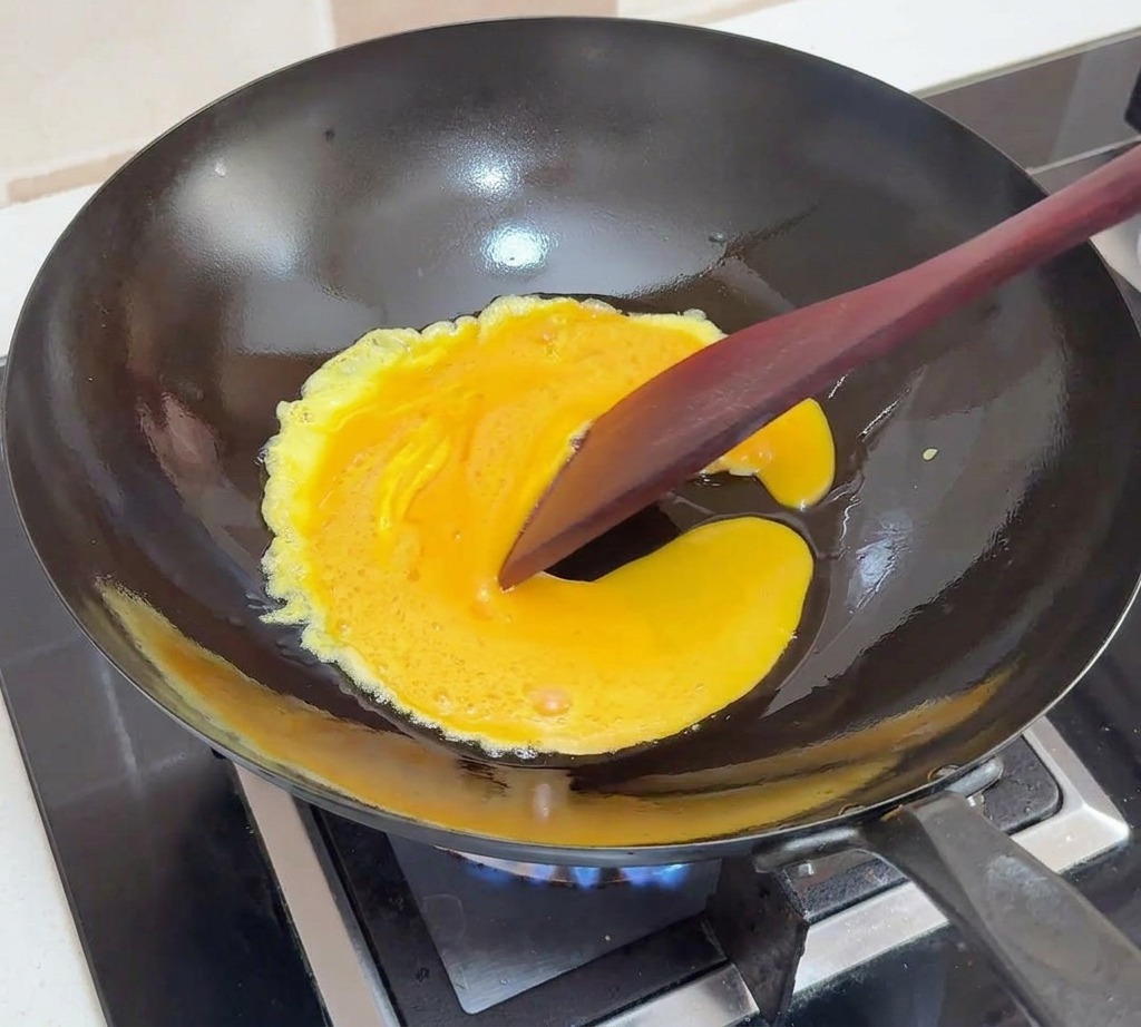 Uova sbattute in cottura in un wok nero, spinte verso il centro con una spatola scura per creare morbide pieghe.