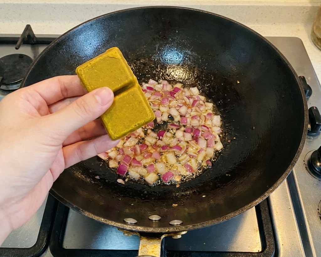 Una persona che tiene due dadi di curry marrone sopra un wok contenente pezzi di cipolla rossa saltati.