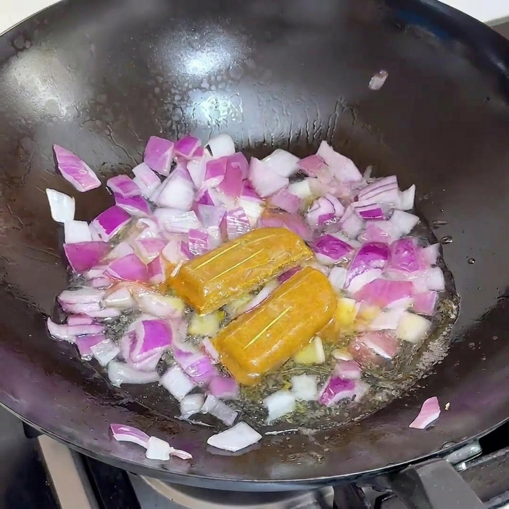 Acqua versata in un wok nero riempito con cipolle rosse e due dadi interi di curry.