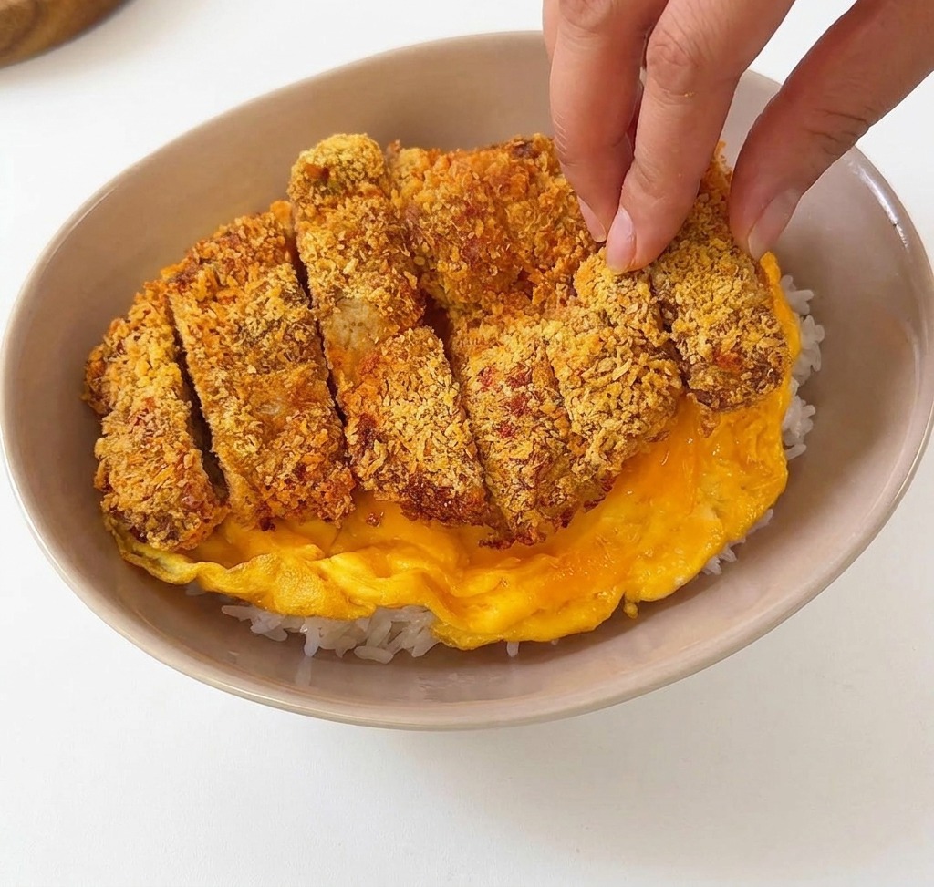 Cotoletta di maiale impanata a fette posizionata sopra una morbida frittata gialla allinterno di una ciotola di ceramica beige con riso bianco.