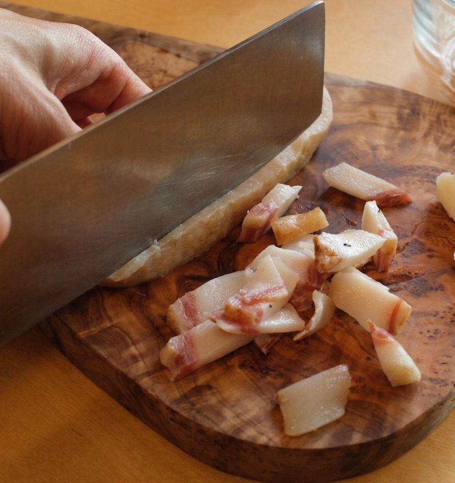Un coltello affilato che rimuove la pelle dura dal guanciale su un tagliere di legno.