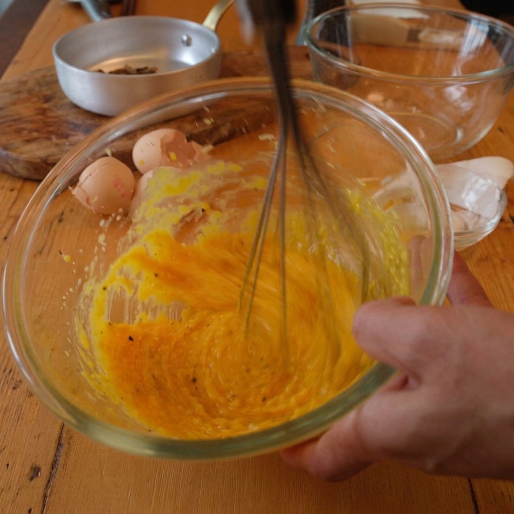 Mano che usa una frusta per unire tuorli e formaggio mentre viene versata acqua calda.