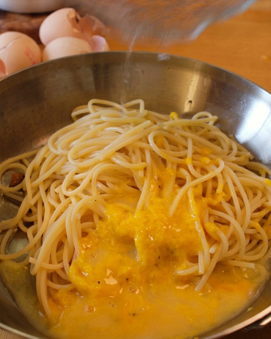 Spaghetti in padella avvolti da una salsa gialla mentre viene aggiunto un filo dacqua di cottura.