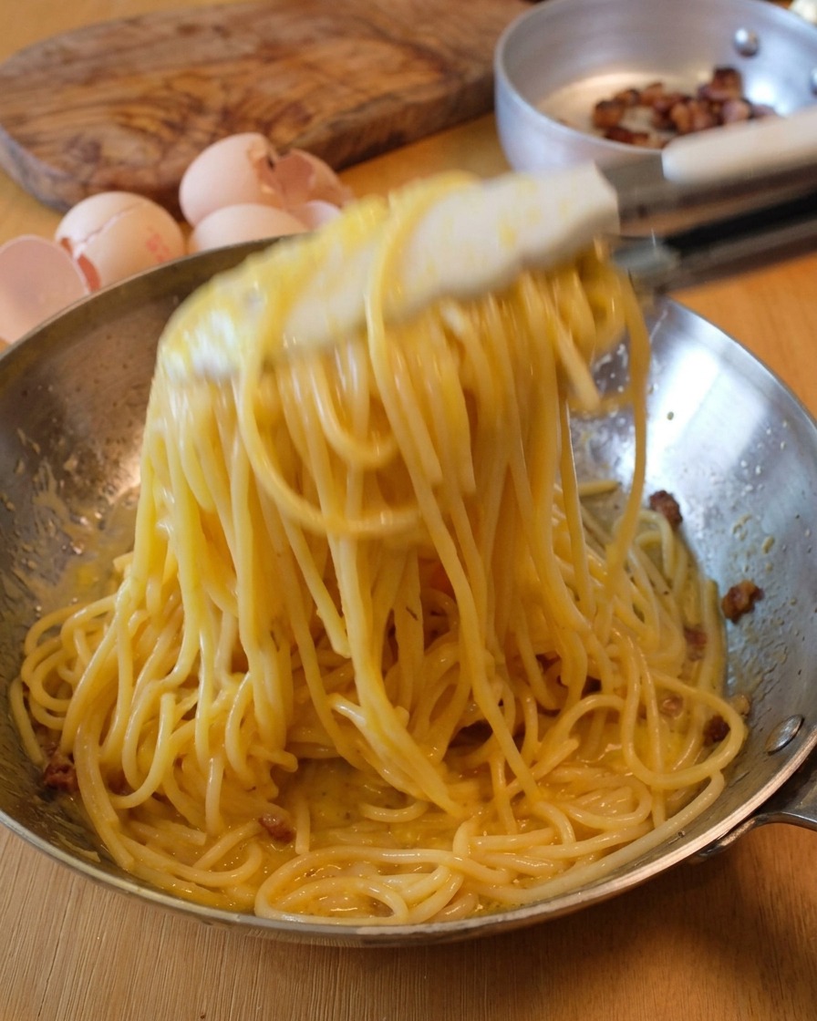 Salto degli spaghetti in padella con pinze per creare la tipica cremosità della Carbonara.