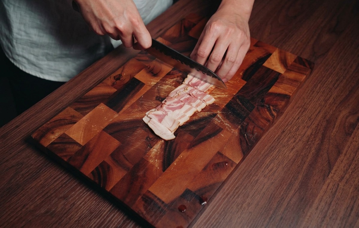 Una persona usa un coltello da chef per tagliare strisce di guanciale a cubetti su un tagliere di legno scuro.