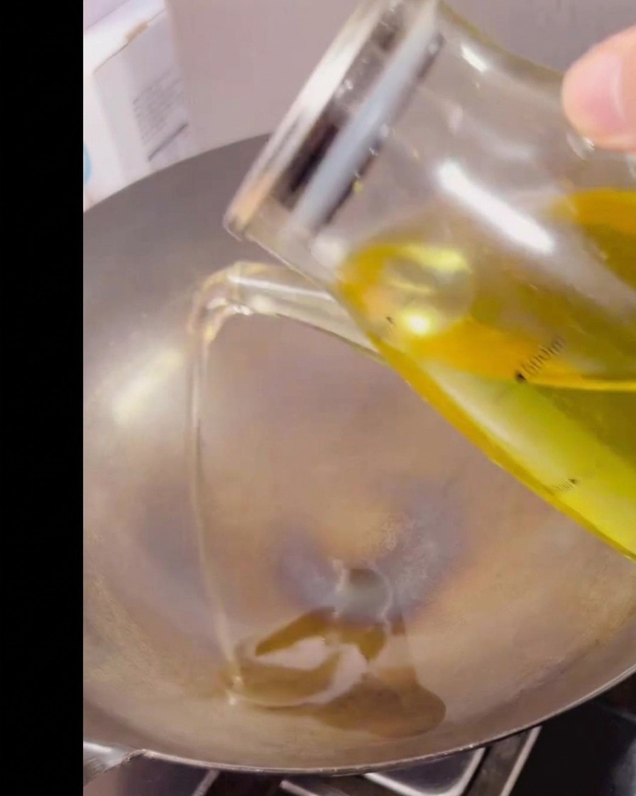 Olio da cucina versato da una caraffa di vetro graduata in un wok di metallo caldo e vuoto.