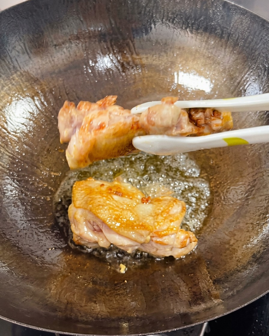 Pinze che girano una sovracoscia di pollo rosolata in un wok, mostrando il lato cotto dorato e croccante.