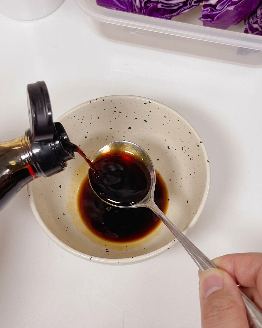 Salsa di soia chiara versata da una bottiglia in un cucchiaio sopra una ciotola contenente salsa scura e condimenti.