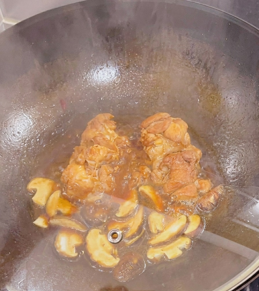 Sovracosce di pollo e funghi shiitake a fette che cuociono a fuoco lento in una salsa scura allinterno di un wok sotto un coperchio di vetro.