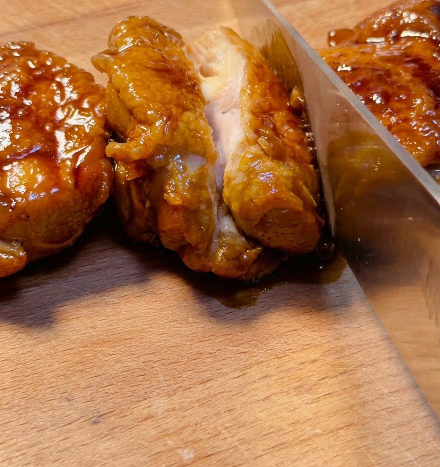 Un coltello affilato che taglia una sovracoscia di pollo teriyaki profondamente glassata su un tagliere di legno, mostrando la carne succosa allinterno.