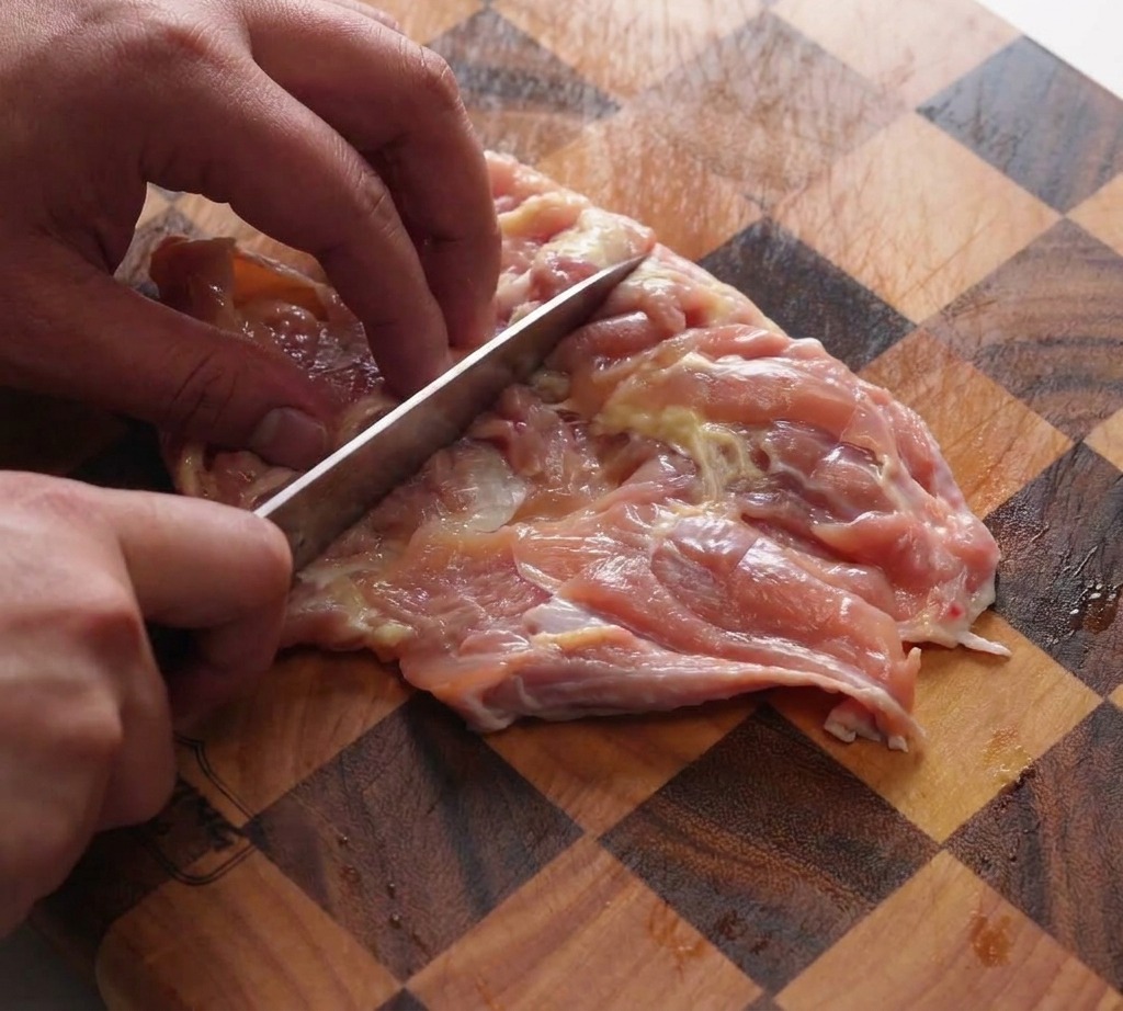 Una mano usa il dorso di un coltello da chef per battere le sovracosce di pollo disossate su un tagliere di legno a scacchi.