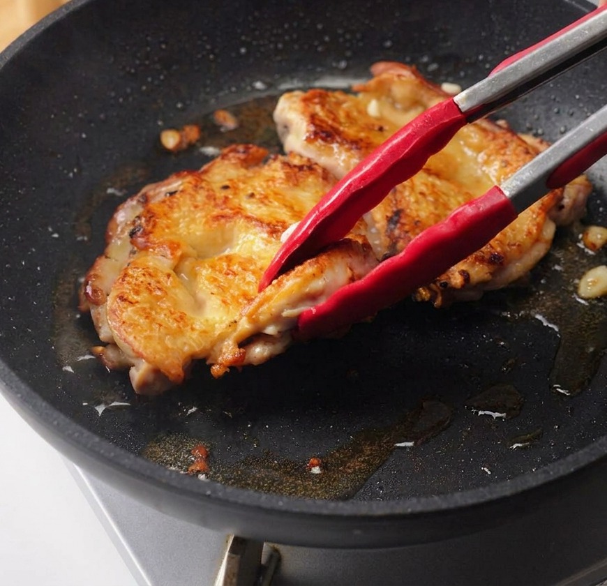 Una mano versa la salsa teriyaki scura da una ciotola di vetro sopra le sovracosce di pollo rosolate in padella.