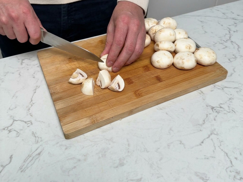Mani che usano un coltello da chef per tagliare i funghi champignon bianchi in quarti su un tagliere di legno.