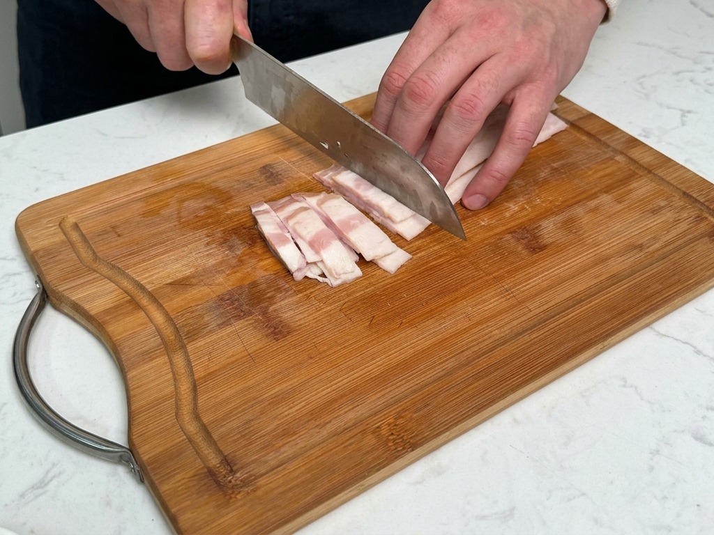 Una persona che taglia a strisce spesse la pancetta su un tagliere di legno.
