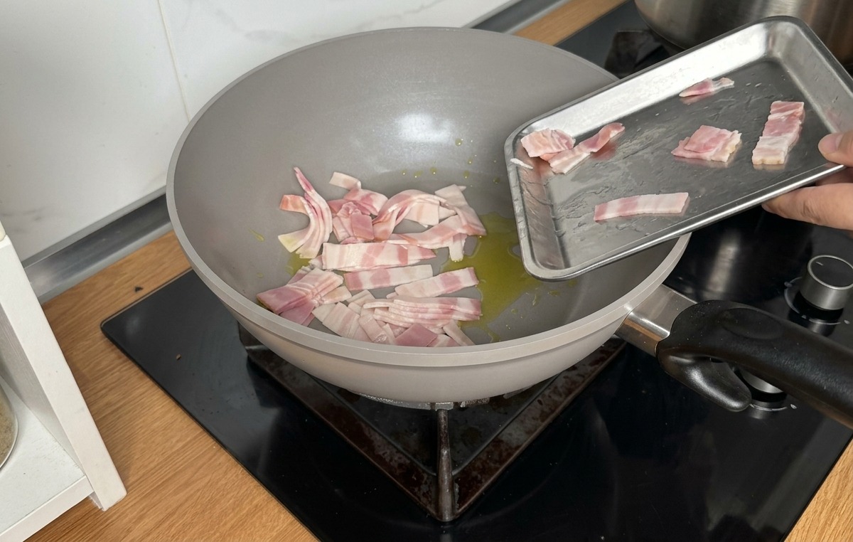Strisce di pancetta cruda fatte scivolare da un vassoio di metallo in una padella grigia con olio doliva.