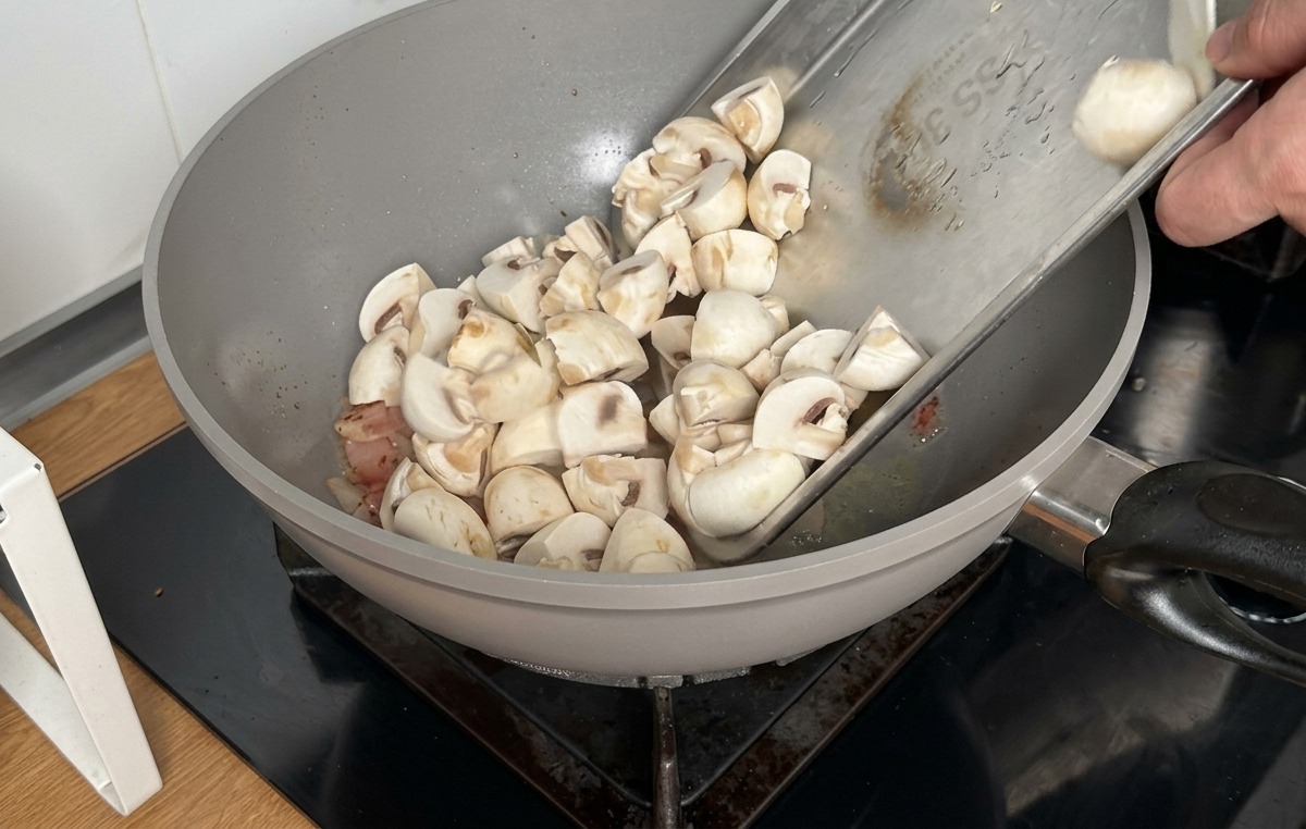 Funghi champignon tagliati in quarti versati da un vassoio di metallo in un wok con la pancetta cotta.