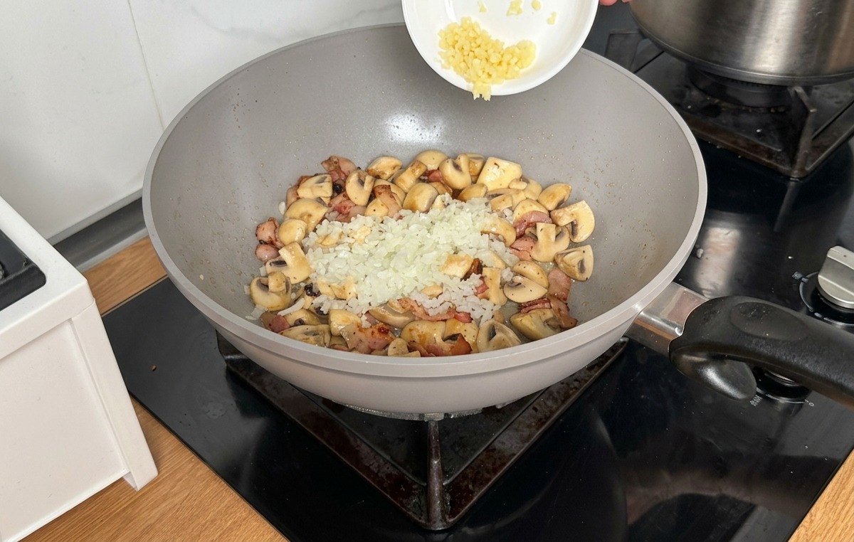 Cipolla e aglio tritati finemente versati da una ciotolina sopra i funghi e la pancetta nel wok.
