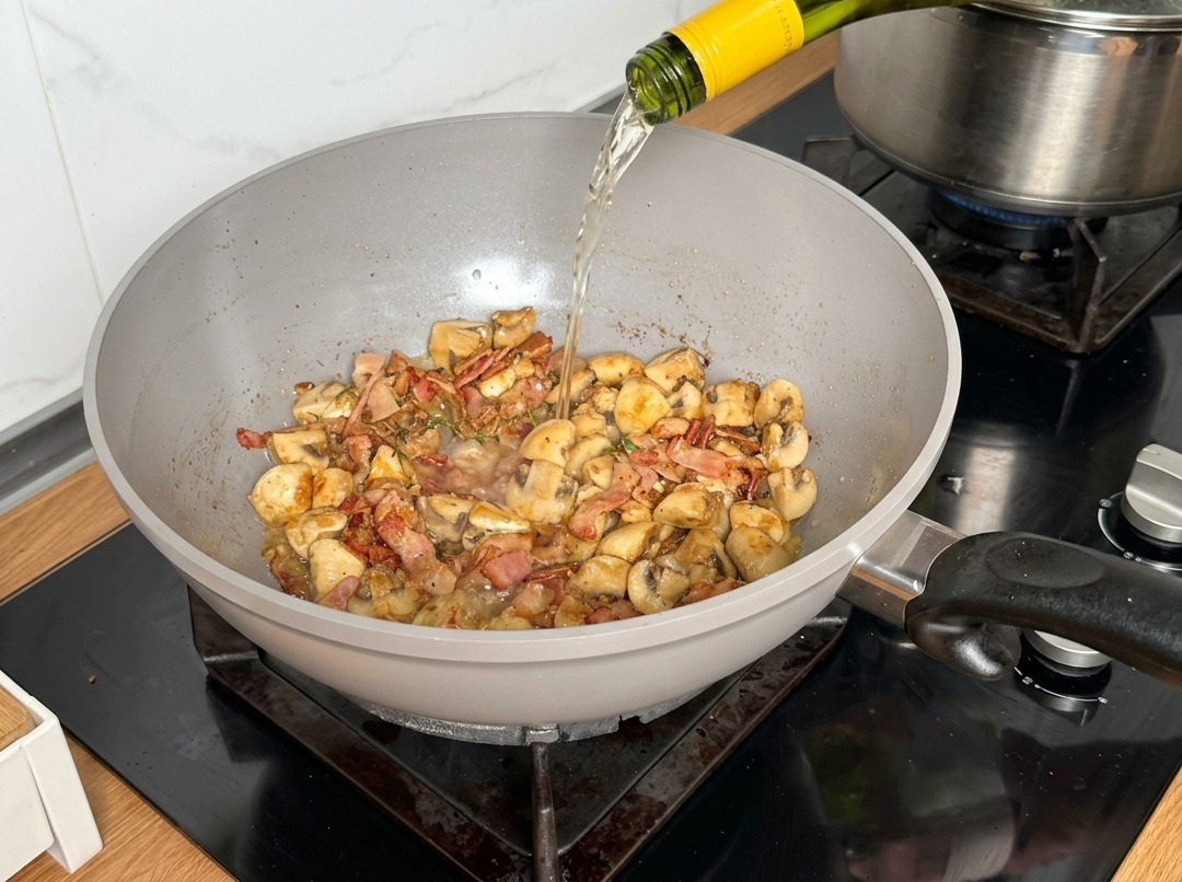 Vino bianco versato da una bottiglia in una padella con funghi e pancetta.