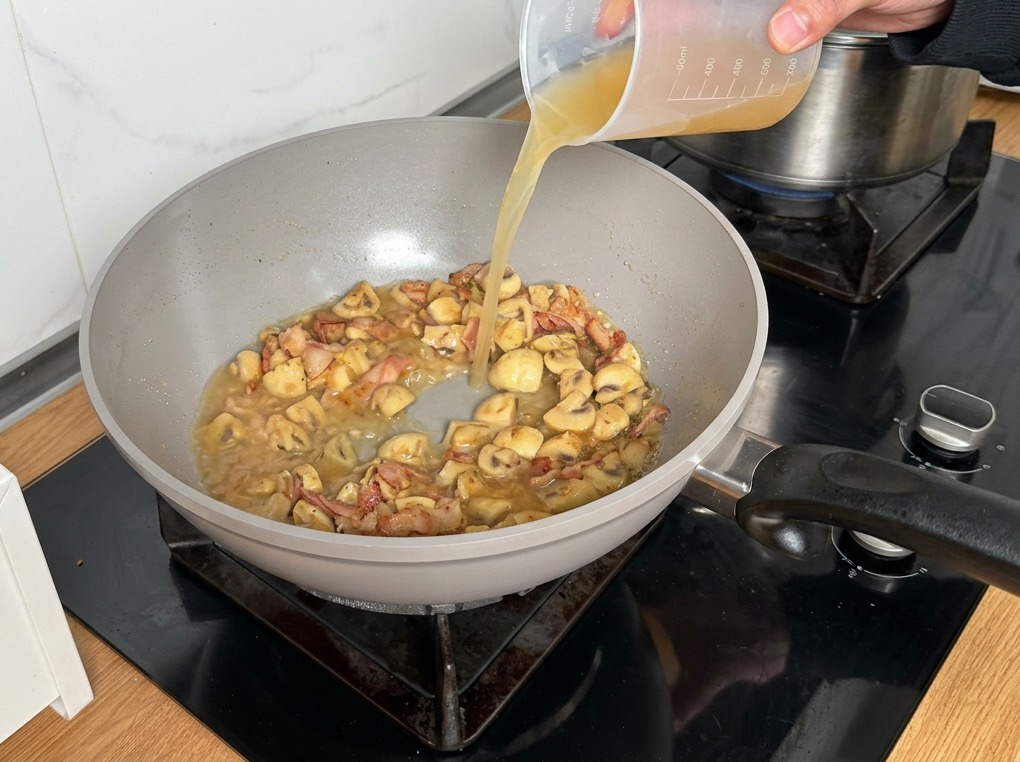Brodo di pollo versato da un misurino di plastica nel mix di funghi e pancetta.