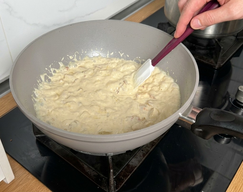 Una spatola bianca che mescola una salsa alla panna densa e bollente con pancetta e funghi.