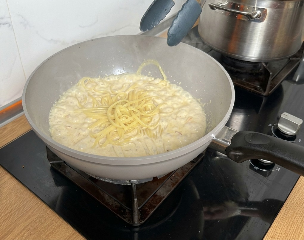 Pinze di metallo che sollevano la pasta cotta dallacqua bollente verso la padella con la crema.
