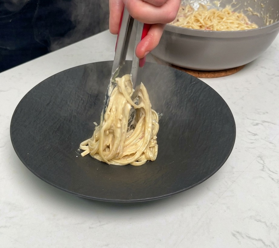 Uso di pinze in metallo per arrotolare e servire una porzione di pasta cremosa su un piatto nero opaco.