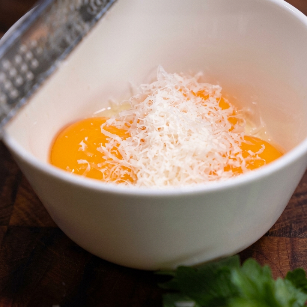 Una grattugia in metallo che fa cadere dei fini riccioli di Parmigiano in una ciotola con tre tuorli crudi.