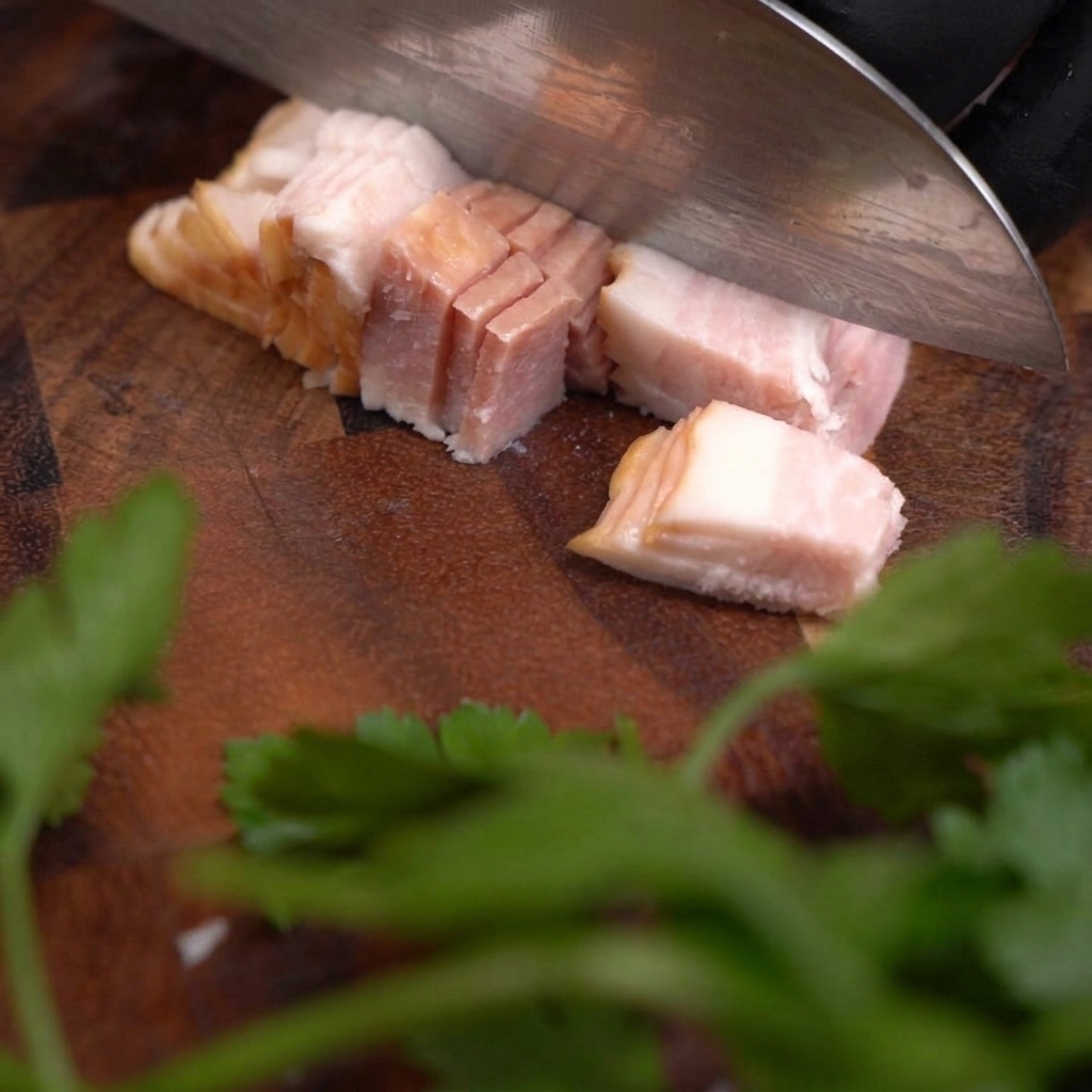 Un coltello affilato che taglia un pezzo di bacon crudo ricavando piccoli cubetti uniformi su un tagliere di legno scuro.