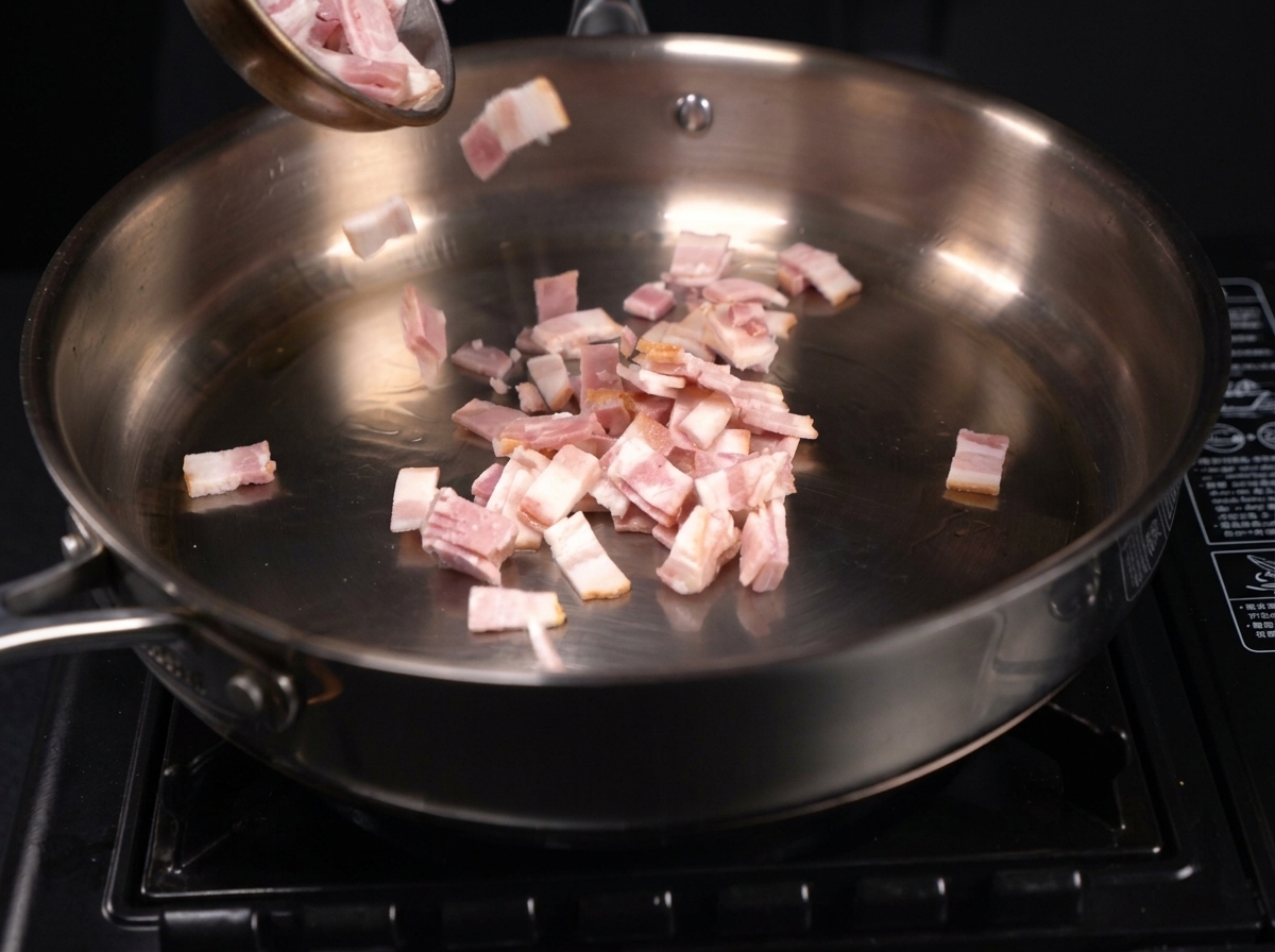 Piccoli pezzi di bacon tagliati che sfrigolano e rilasciano il loro grasso in una padella di acciaio inossidabile.