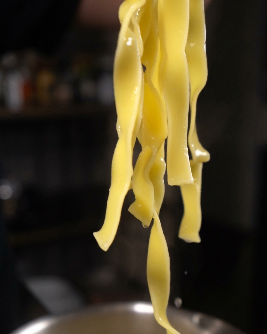 Lunghe e spesse strisce di tagliatelle gialle cotte appese a delle pinze, dalle quali gocciola dellacqua.