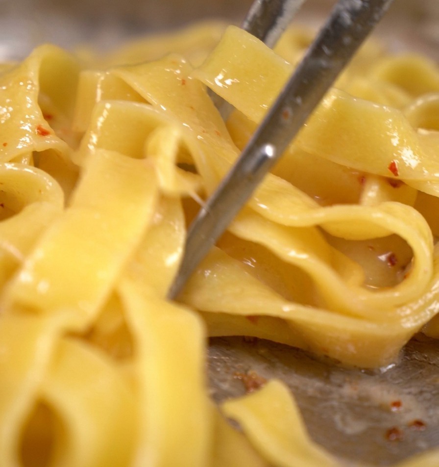 Tagliatelle saltate con pinze di metallo in una padella con pezzetti di bacon e acqua di cottura della pasta che sobbolle.