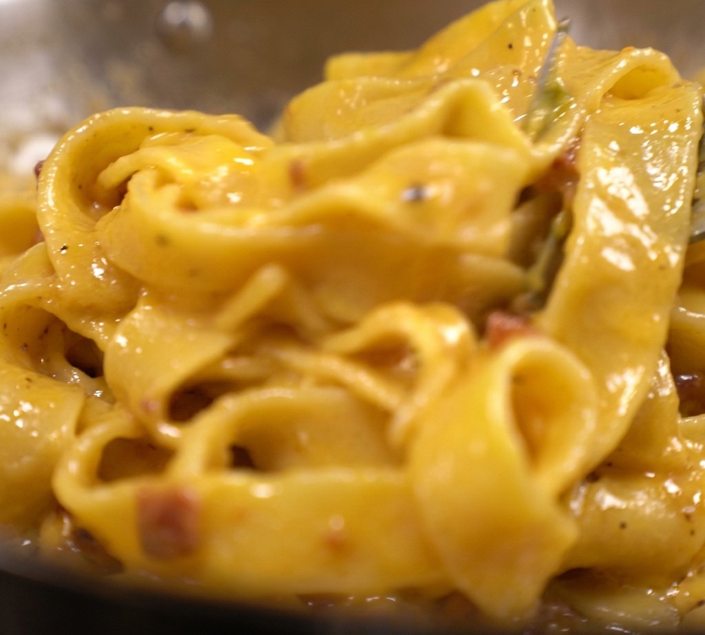 Tagliatelle ricoperte da una spessa e vibrante crema gialla a base di uovo e formaggio, con visibili pezzi di bacon in una padella.