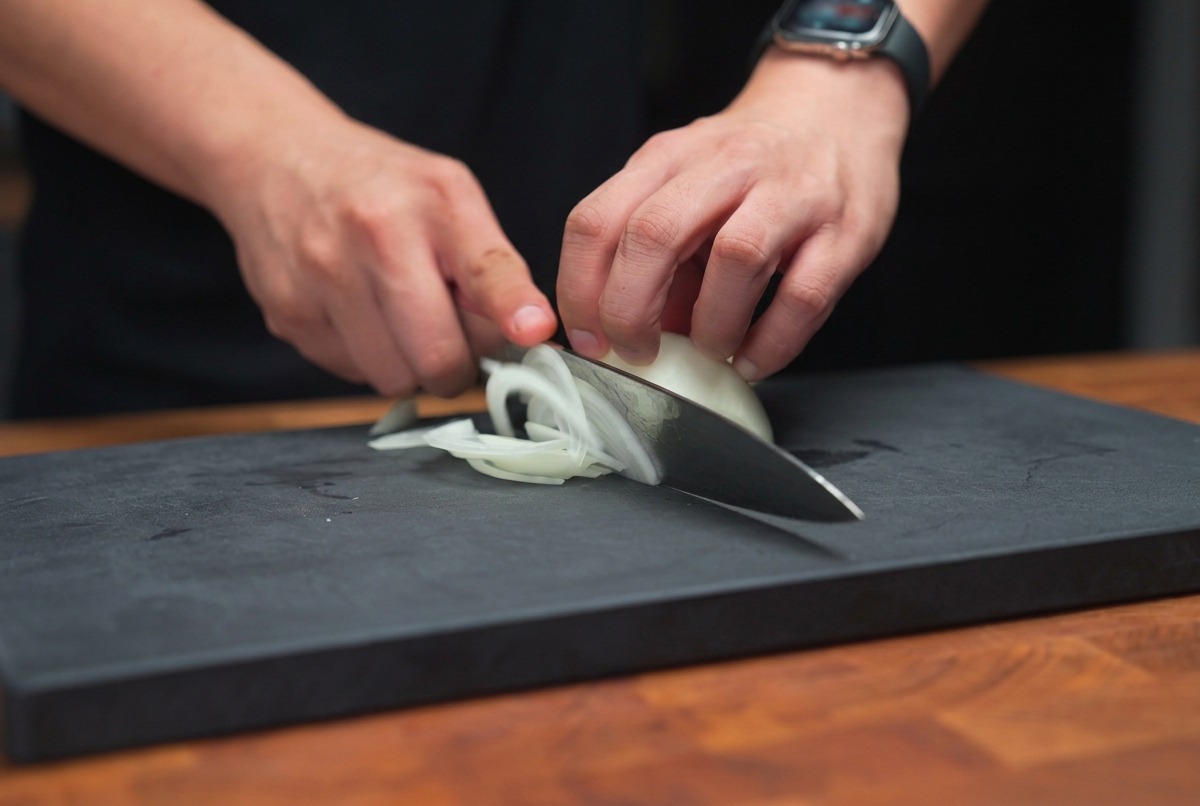 Mani che impugnano un coltello da chef e tagliano una cipolla bianca a listarelle sottili su un tagliere scuro.