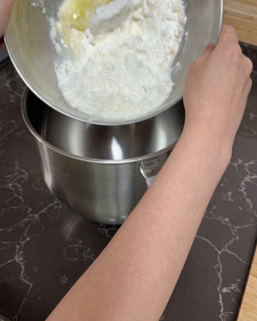 Mani che versano un composto grezzo di farina e ingredienti liquidi nella ciotola vuota di una planetaria.