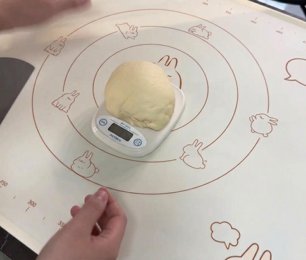 Una grande palla di impasto rotonda e liscia appoggiata su una bilancia da cucina digitale bianca sopra un tappetino in silicone per cottura.