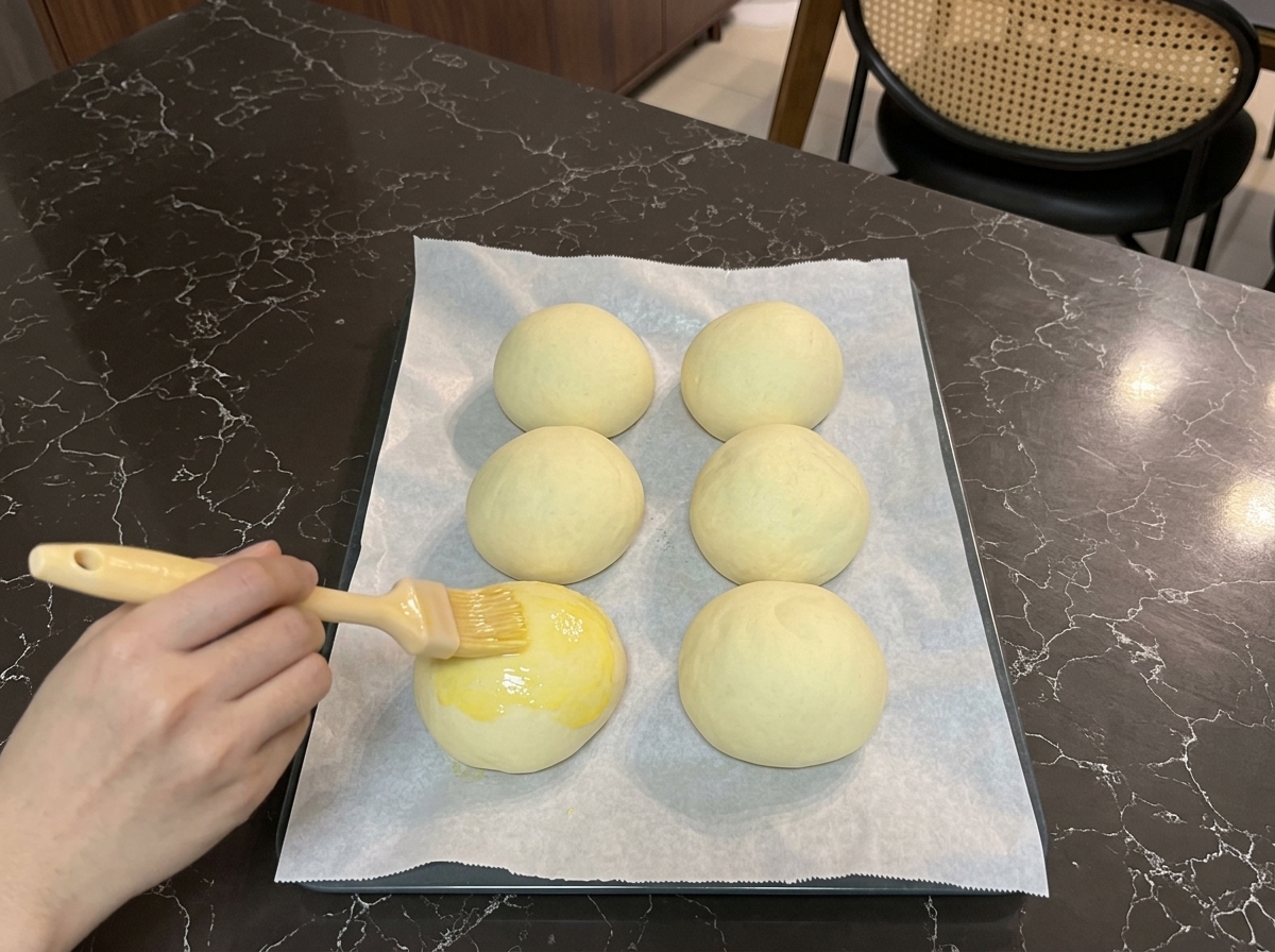 Una mano che usa un pennello da cucina per ricoprire la parte superiore di un panino lievitato con una lucida miscela gialla di uovo.